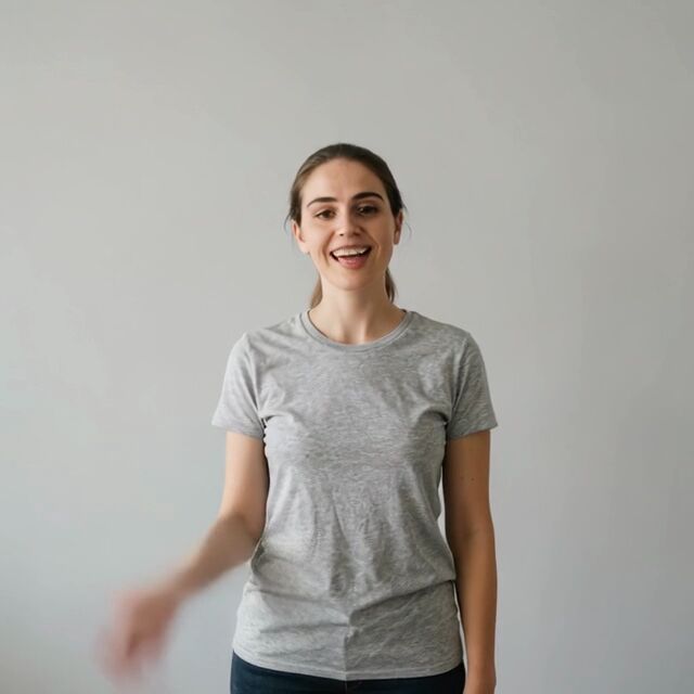 A woman in a grey t-shirt against a white wall, waving and speaking to camera