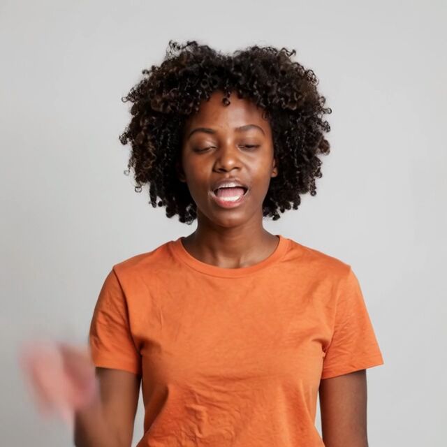 A different woman, in an orange t-shirt, making the same wave and speaking the same words